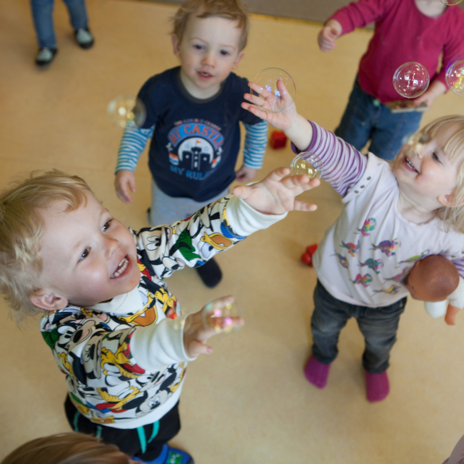 Kleine Kinder spielen mit Seifenblasen und reißen die Arme hoch.