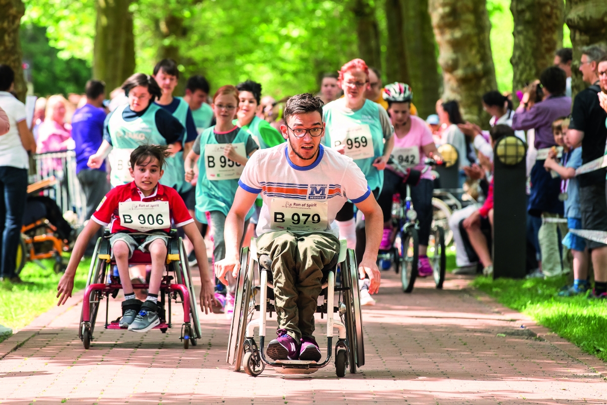 Ein Jugendlicher und ein Kind im Rollstuhl beim Run of Spirit. Dahinter befinden sich weitere Läuferinnen und Läufer mit und ohne Behinderung.