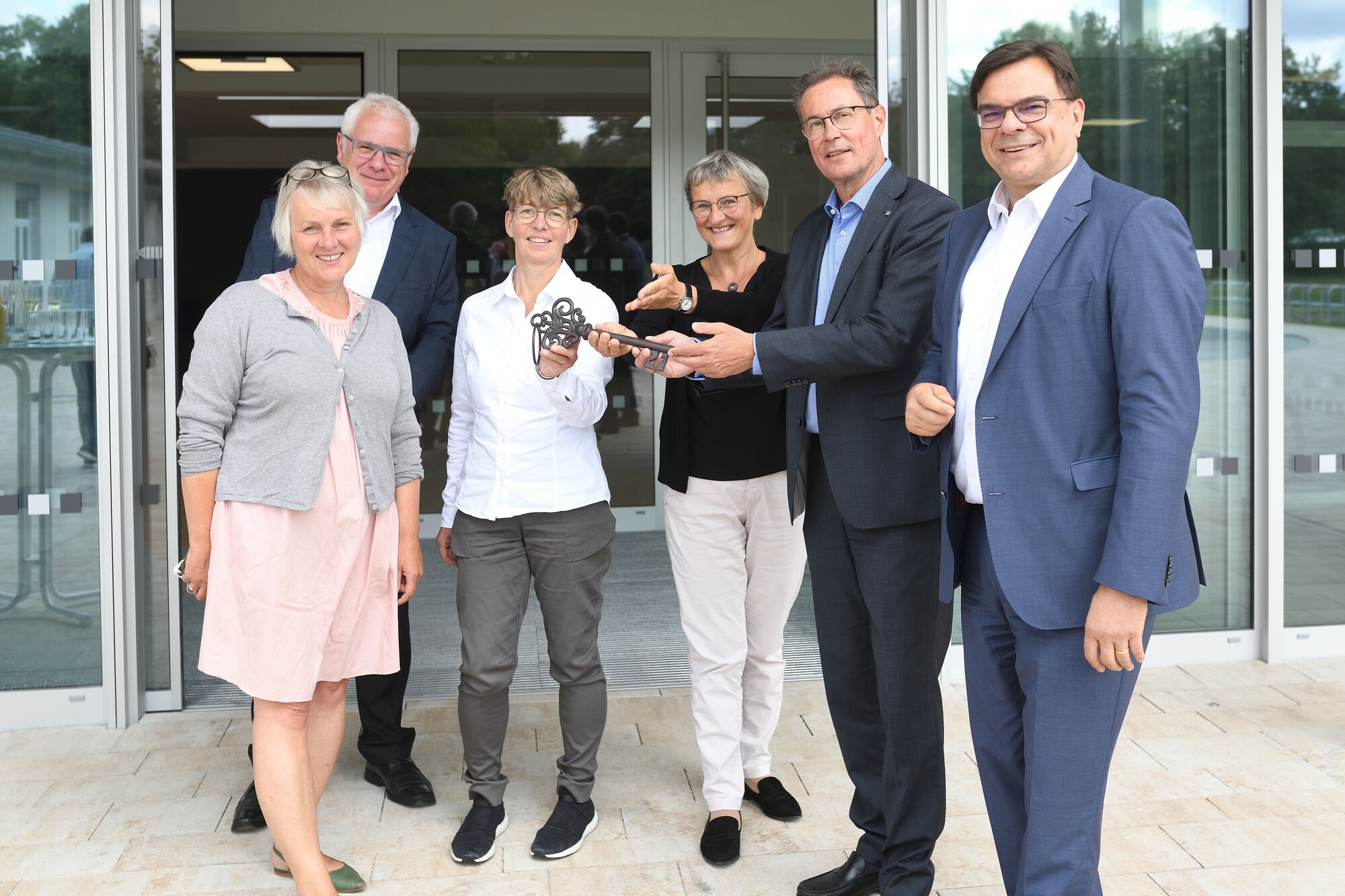 Mehrere Menschen stehen nebeneinander vor dem neuen Schulgebäude. Eine Mann und eine Frau halten zur symbolischen Übergabe einen Schlüssel in die Höhe.